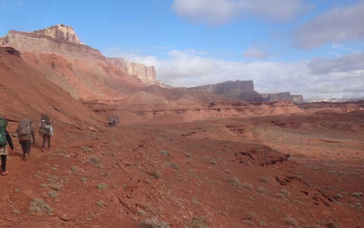 A group of students wearing backpacks hike along a trail in a red desert landscape